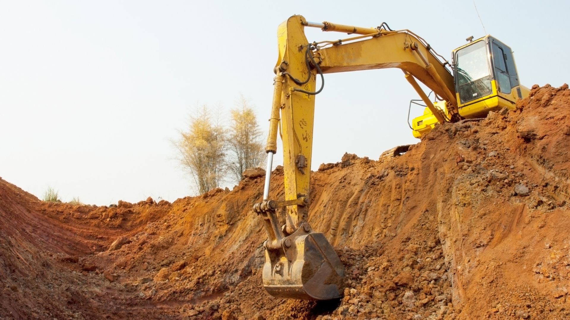 Yellow excavator digging in reddish-brown soil with trees in background