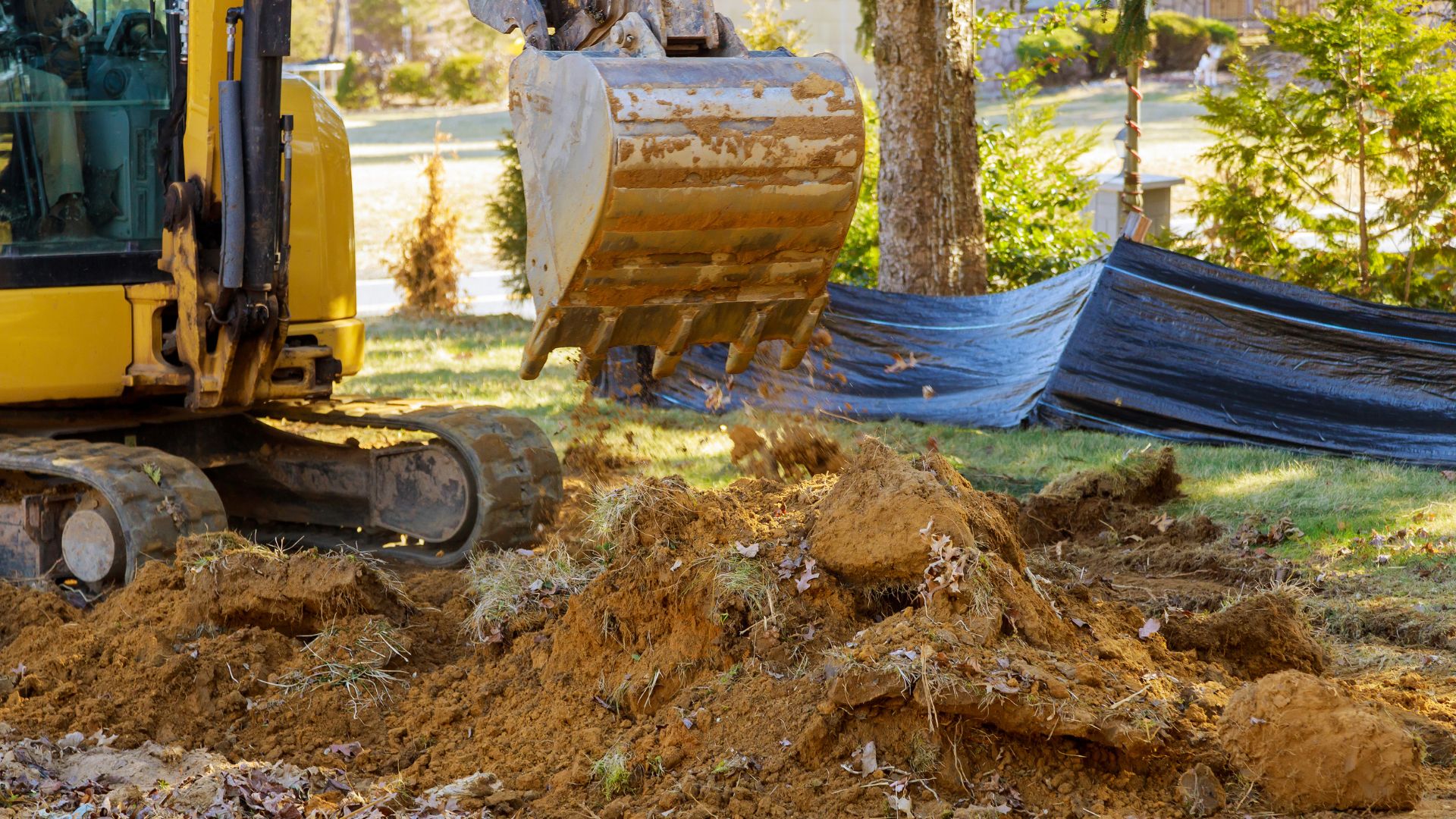 Yellow excavator digging up dirt in a yard with blue tarp nearby